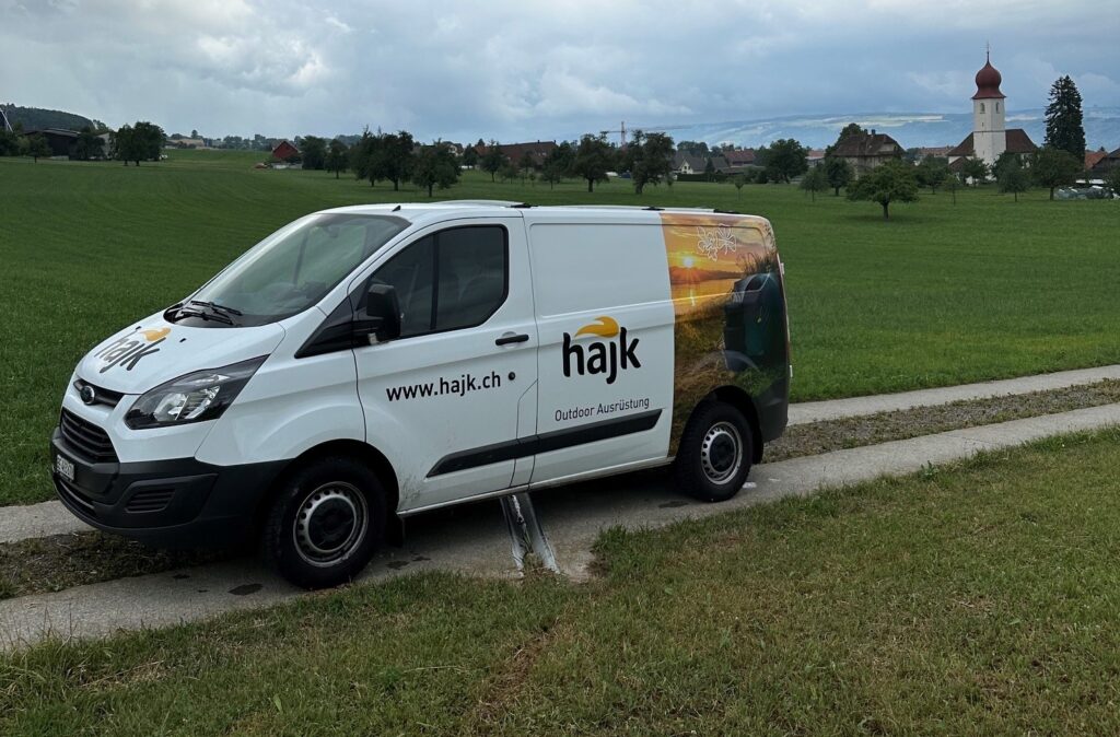 Weisser hajk-Lieferwagen mit Firmenlogo und der Aufschrift www.hajk.ch auf einem Feldweg in einer verlassenen Wiese vor einer Dorfkirche.