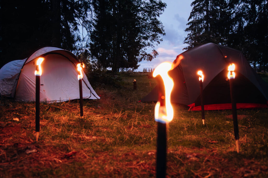 Brennende Wachsfackeln stecken im Waldboden und beleuchten in der Abenddämmerung eine Lichtung, auf welcher zwei kleine Zelte aufgebaut sind.