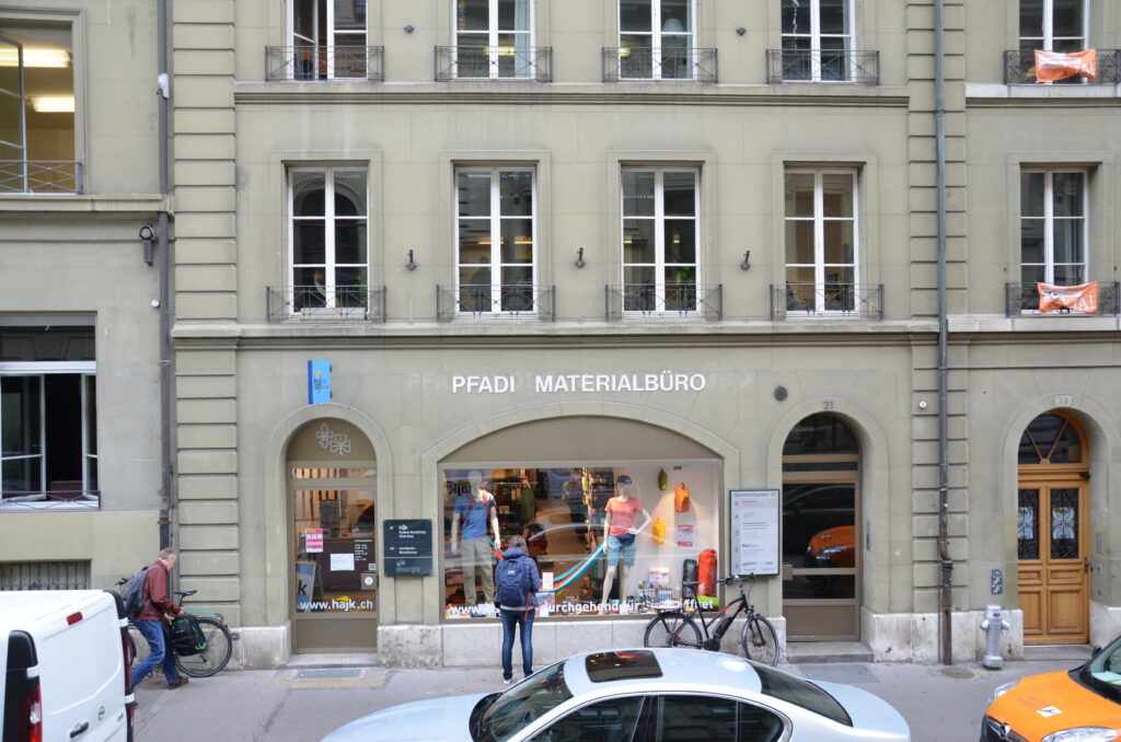 Frontansicht des heutigen hajk-Shops an der Speichergasse 32 in Bern in Farbe. Vor dem Haupteingang sind diverse Autos parkiert.