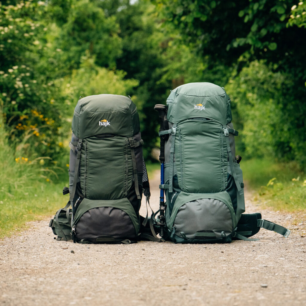 Zwei hajk-Scoutpack-Rucksäcke stehen aufgestellt auf einer Naturstrasse, die durch einen Wald führt.